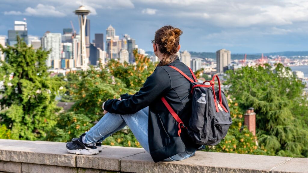 Young lady backpacker tourist alike visiting Queen Anne Hill's Kerry Park to catch a spring sunset over downtown Seattle and Mt. Rainier in the background.